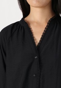 Svart skjorta med knappar i en strukturerad fabric, med en V-ringning prydd med spets och insydda axeldetaljer. Knapparna är svarta.