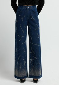 Jeans de mezclilla azul de pierna ancha con adornos metálicos que forman un patrón en espiral. Textura suave con un ligero efecto de desgaste en los puños.