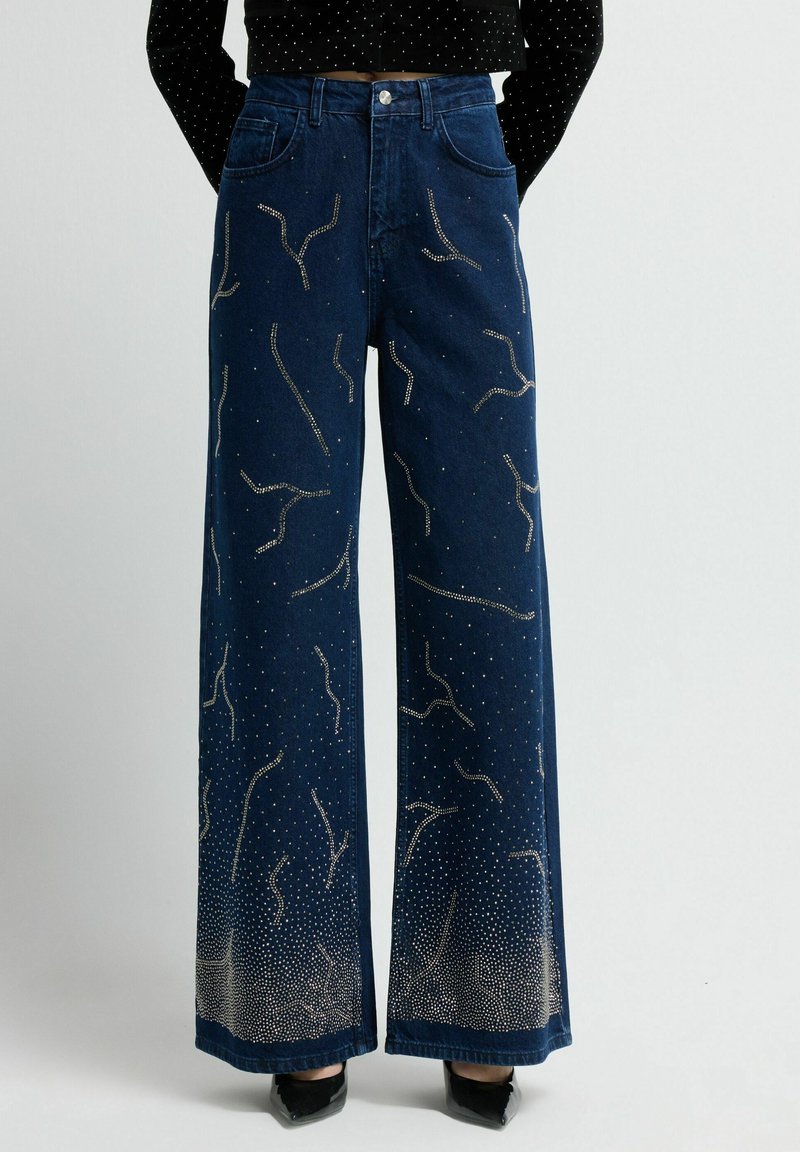 Jeans de mezclilla azul de pierna ancha con adornos metálicos que forman un patrón en espiral. Textura suave con un ligero efecto de desgaste en los puños.