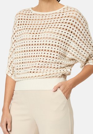 Femme portant un pull beige à maille lâche par-dessus un top blanc, une main dans la poche d'un pantalon beige clair sur un fond uni.