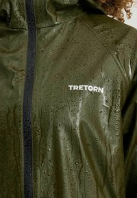 Vattentät grön regnjacka med en glänsande yta, med en svart dragkedja och varumärket "TRETORN" i vit text. Vattendroppar kan ses på ytan.
