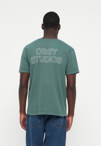 Grøn bomulds T-shirt med rund hals, med teksten "OBEY STUDIOS" trykt på ryggen i hvid, kortærmet og med en afslappet pasform.