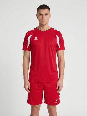 Jeune homme debout, faisant face, portant un maillot de sport rouge et un short assorti avec des détails en chevrons blancs sur les manches et les côtés.