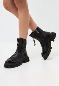 Botine negre cu accente lucioase din patent, piele texturată, talpă groasă și fermoare laterale pentru o purtare ușoară.