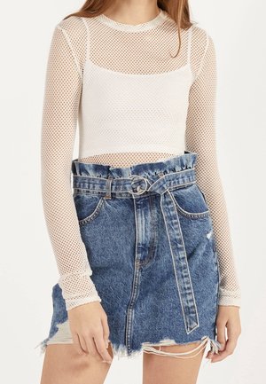 Mujer con top corto de malla blanca de manga larga y falda vaquera azul de talle alto con cintura fruncida y cinturón con hebilla de doble anilla.