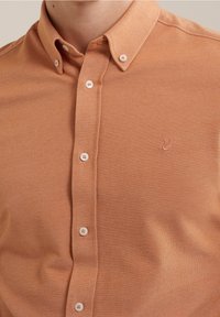 Světle oranžová košile na knoflíky s texturovaným materiálem, má límec, bílé knoflíky a malou vyšitou nášivku na hrudi.