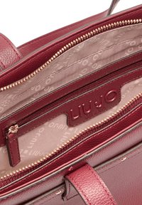 Interior de bolso de cuero burdeos con un forro de tela claro, que incluye un bolsillo con cremallera, cremalleras en tono dorado y logo de la marca en relieve.