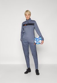 Sivi sportski jakna s crnim detaljima i ovratnikom na zatvarač, uparen s odgovarajućim sivim hlačama. Crne sportske cipele i plavi nogometni lopta.