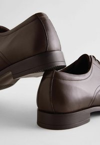 Zapatos de cuero marrón con una textura suave, forma redondeada, detalles de costura sutiles y un tacón bajo de goma.
