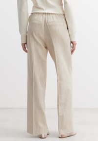Beige weit geschnittene Hosen aus strukturiertem Stoff, mit elastischem Taillengummi und zwei Gesäßtaschen. Getragen mit einfachen Riemchensandalen.