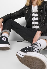 Chaqueta de moto de cuero negra, camiseta gráfica blanca, jeans negros con los dobladillos enrollados, zapatillas Converse negras y calcetines blancos sobre un suelo gris.