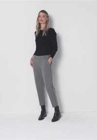 Vrouw staand tegen een effen muur, gekleed in een zwarte top met lange mouwen, grijze op maat gemaakte broek en zwarte enkellaarsjes, met handen in de zakken.