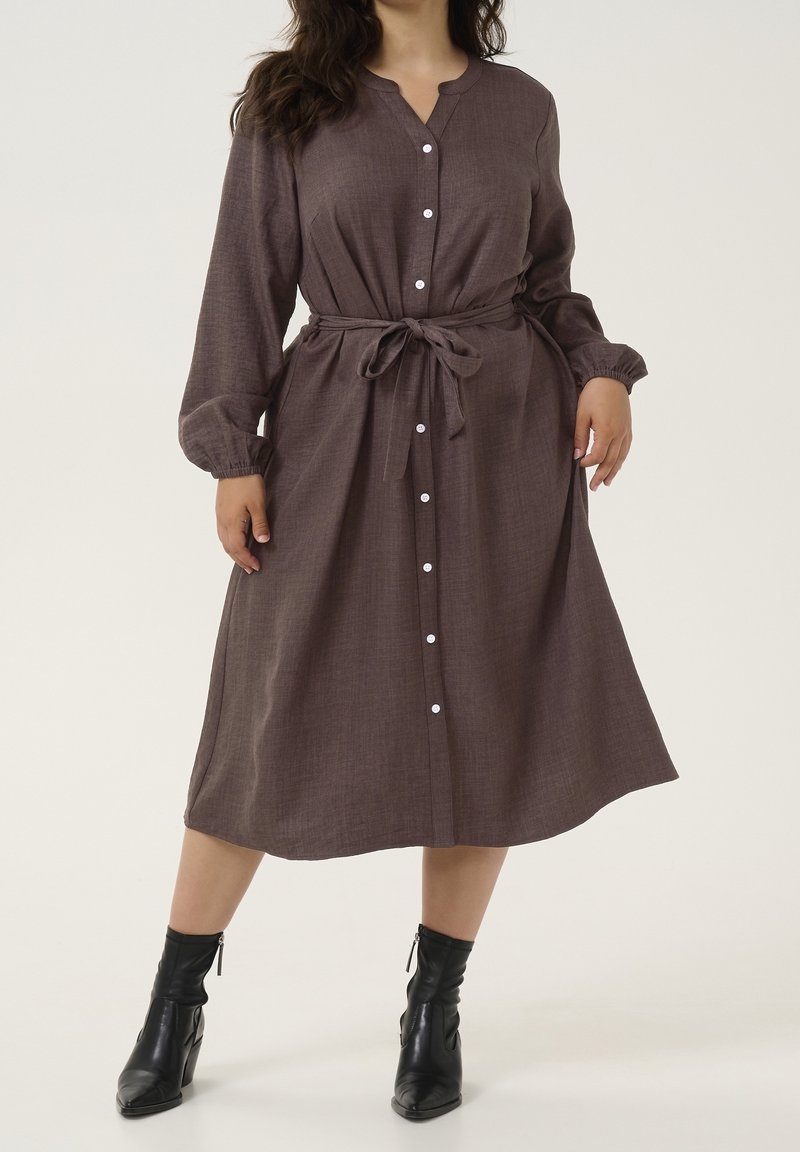 Femme portant une robe midi marron à manches longues boutonnée, avec une ceinture à la taille et des bottines noires, sur un fond uni.