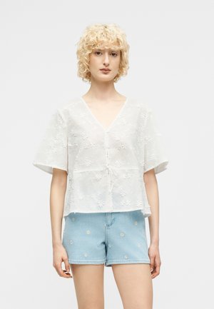 Młoda osoba o kręconych blond włosach, ubrana w białą kwiatową koszulę na guziki oraz jasnoniebieskie krótkie spodenki dżinsowe z małymi białymi nadrukami stokrotek.