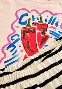 Vaaleanpunainen t-paita, jossa on värikäs chili-paprika -graafinen kuvaus sinisillä ja vihreillä yksityiskohdilla, yhdistettynä musta-valkoisiin raidallisiin joustovyöhön.
