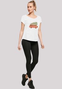 Witte t-shirt met korte mouwen, voorzien van een afbeelding van een vintage rode bus met een kerstboom erop, gecombineerd met zwarte leggings en zwarte sneakers.