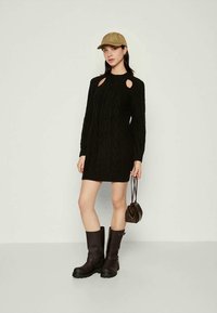 Μαύρο πλεκτό φόρεμα με λεπτομέρειες κοψίματος στους ώμους, μακριά μανίκια και άνετη εφαρμογή· συνδυασμένο με μαύρες μπότες μέχρι τα γόνατα και μπεζ καπέλο.