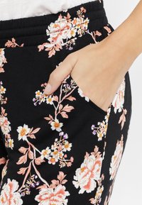 Zwarte broek met bloemenprint, voorzien van een zijzak, elastische tailleband en een patroon van roze en witte bloemen met groene accenten.
