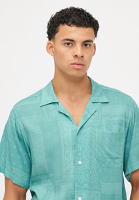 Camisa de manga curta turquoise com um bolso, apresentando um padrão de paisley e carinhas sorridentes por toda a parte, feita de tecido leve.