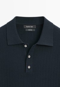 Polo en coton côtelé bleu marine foncé avec trois boutons blancs et un col classique, étiqueté Massimo Dutti, fabriqué en Chine.