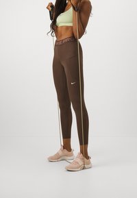 Leggings atletiche marroni con vita alta e tasca laterale, abbinate a un reggiseno sportivo verde chiaro e scarpe da allenamento rosa. Inclusi bande di resistenza.