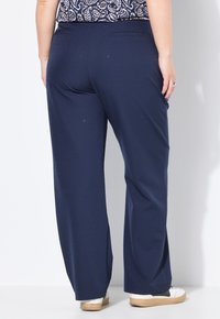 Persona che indossa pantaloni larghi blu navy con una sottile texture a pois, sneakers bianche con suole in gomma, in piedi contro un muro bianco.