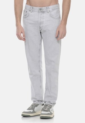 Uomo che indossa jeans grigio chiaro a gamba dritta e sneakers bianche con dettagli grigi, in piedi davanti a uno sfondo bianco uniforme.