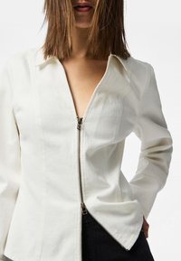 Camicia bianca a maniche lunghe con scollatura a V e chiusura con zip obliqua; presenta un design aderente e una texture liscia.