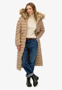 Lang beige pufferjakke med hætte med pelskant, quiltet tekstur og lynlås-lommer i siden, båret over en lys sweater og blå jeans.