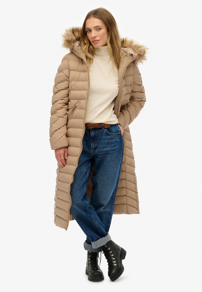 Lang beige pufferjakke med hætte med pelskant, quiltet tekstur og lynlås-lommer i siden, båret over en lys sweater og blå jeans.