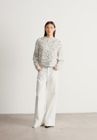 Grober Strickpullover in Hellgrau mit bunten Farbtupfern, kombiniert mit weißen Hosen mit weitem Bein, die eine hohe Taille und klare Linien aufweisen.