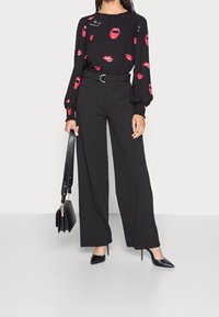 Kvinna iförd svarta vida byxor och en svart blus med röda läppmönster, hållande en svart handväska och bärande svarta spetsiga klackar.