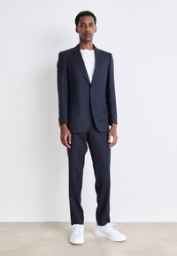 Completo blu navy realizzato in tessuto testurizzato, con giacca sartoriale a due bottoni e pantaloni con frontale piatto, abbinato a sneaker bianche.