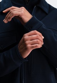 Marineblauwe jas met ritssluiting, knoopmanchetten en platte zakken. Zachte textuur en gestructureerde kraag. Close-up van handen die de mouw aanpassen.