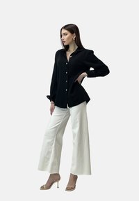 Camisa negra de botones con una tela texturizada, pantalones blancos de pierna ancha y sandalias de tacón nude. La modelo posa de lado contra un fondo claro.