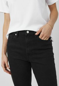Jean en denim noir à taille haute, design à cinq poches, détails de boutons en argent et légèrement extensible. Associé à un t-shirt blanc.