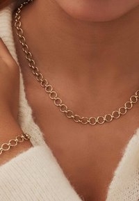 Goldkette mit kreisförmigen Gliedern, die eine glatte Textur aufweist. Trägt man um einen nackten Hals, vor einem hellfarbigen Pullover.