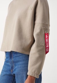 Sweat-shirt court beige en tissu doux avec un bord-côte au poignet. Dispose d'une poche zippée sur la manche ornée d'une étiquette rouge "REMOVE BEFORE FLIGHT".