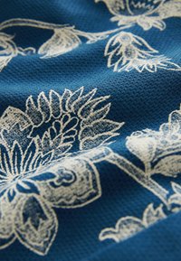 Tessuto blu con superficie testurizzata che presenta un motivo floreale bianco, comprendente fiori grandi e piccoli, con dettagli intricati su tutta la superficie.