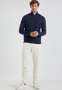 Marineblauwe gebreide trui met een ritssluiting aan de hals, lange mouwen en een gestructureerde afwerking, gecombineerd met lichtgekleurde broeken en witte sneakers.