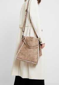 Beige skuldertaske i kunstig ruskind med en tekstureret finish og hvide detaljer, udstyret med en lang rem og en karakteristisk sidekvast.