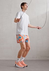 Bela kratka športna majica s kratkimi rokavi, večbarvne kratke hlače v oranžni in modri barvi ter roza športni čevlji. Vrv za skipping je v gibanju.