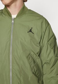 Giacca bomber verde oliva con chiusura a zip, colletto a coste e logo ricamato nero sul petto, realizzata in un tessuto di nylon texturizzato.