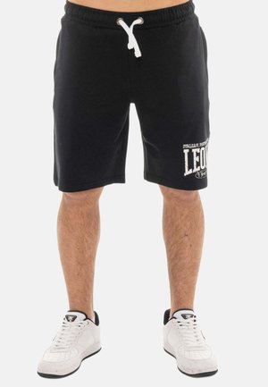 Uomo che indossa pantaloncini neri con coulisse e scritta bianca "LEONE" sulla gamba sinistra e sneakers bianche con dettagli neri, in piedi su sfondo semplice.