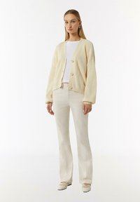 Cremfarbener gerippter Cardigan mit vier Knöpfen, getragen über einem weißen Oberteil, kombiniert mit hellbeigen Schlaghosen und cremfarbenen Loafers. Insgesamt glatte Textur.
