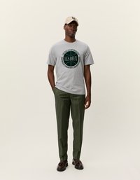 Sivý tričko s zeleným grafickým logom, skombinované so zelenobielymi pruhovanými nohavicami a béžovou čiapkou, nosené so tmavými topánkami.