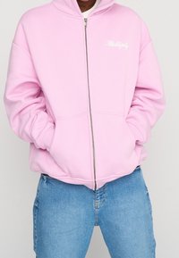 Haut à zip rose en matériau doux, avec une fermeture éclair à l'avant et des poches kangourou. Logo "Multiply" brodé en blanc sur le côté gauche de la poitrine.