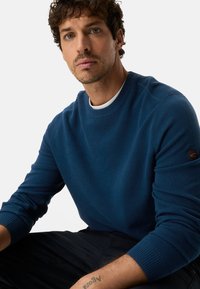 Blue knit sweatshirt featuring a round neckline, ribbed cuffs, and a textured finish, with a logo patch on the left sleeve.