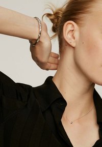 Collar de plata con un pequeño colgante redondo, usado con una camisa de rayas negras. La pulsera en la muñeca presenta un diseño de eslabones.