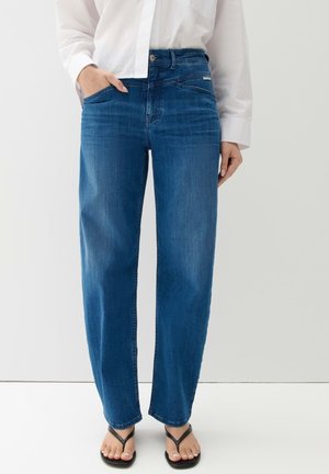 Personne portant un jean bleu droit, une chemise blanche boutonnée et des tongs noires, debout avec une main dans la poche.